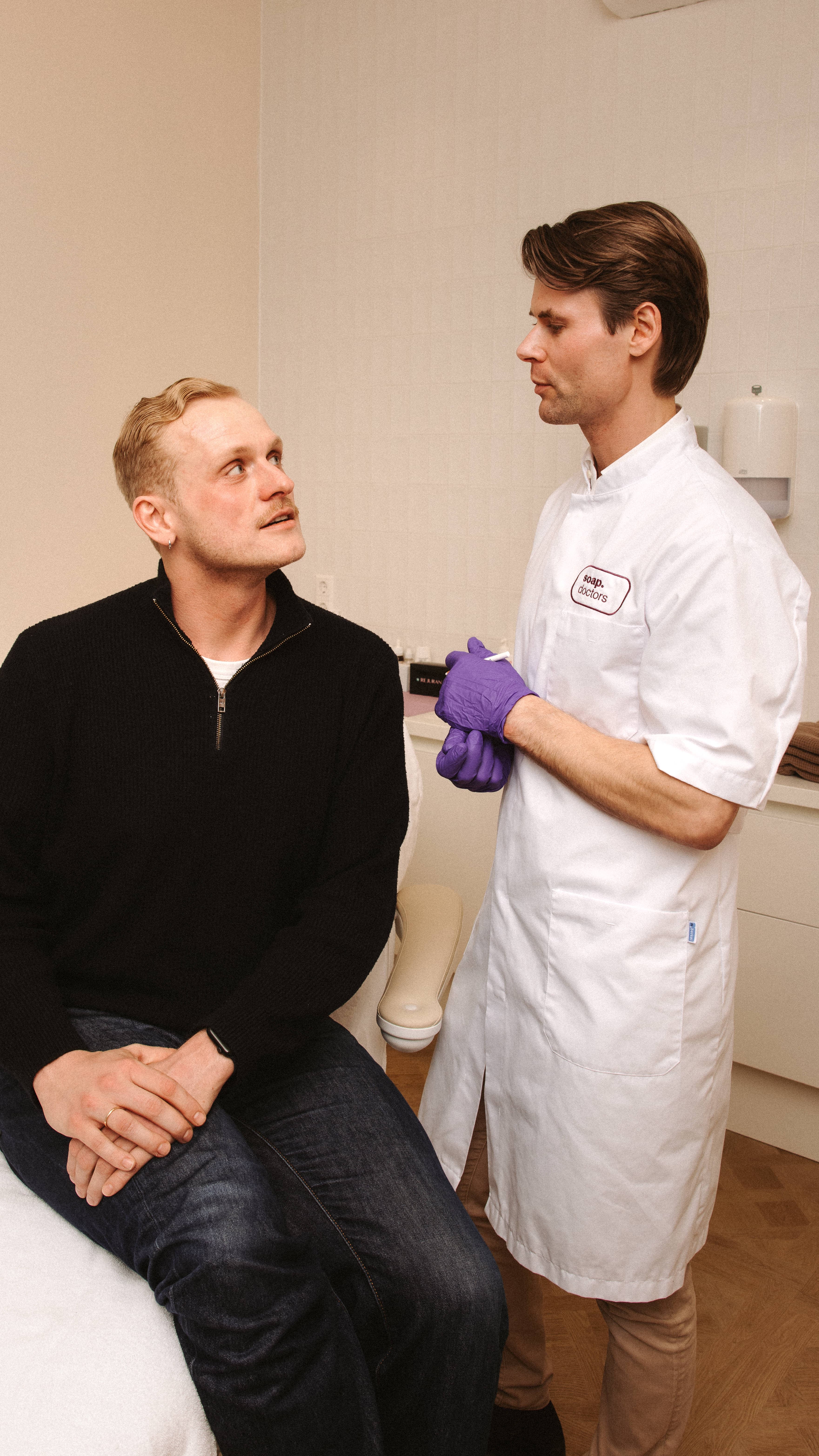 A doctor wearing a white coat and purple gloves consults with a seated patient in a medical office.