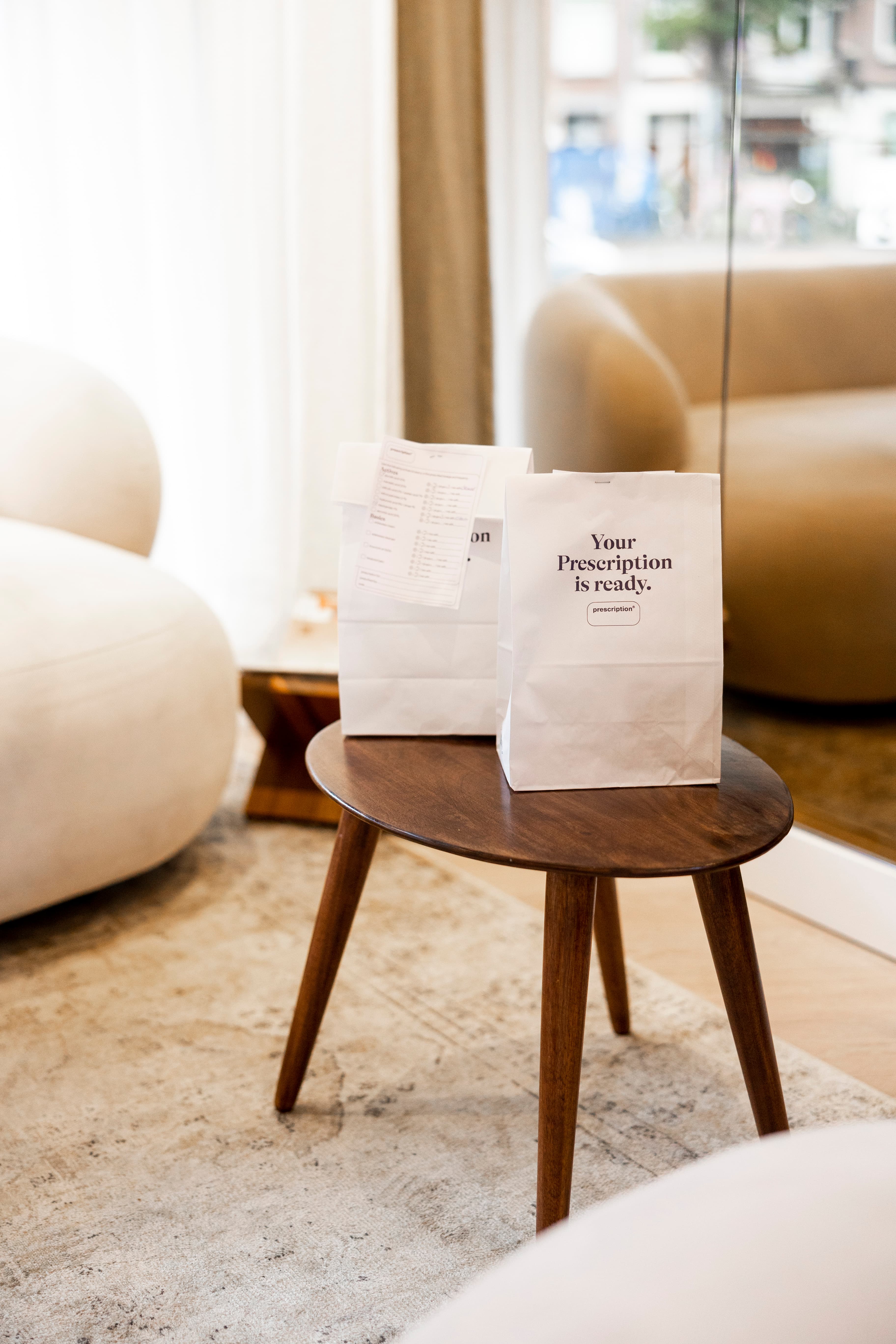 Two white paper bags with "Your Prescription is ready" text on a small wooden table, set in a modern, cozy living room space.