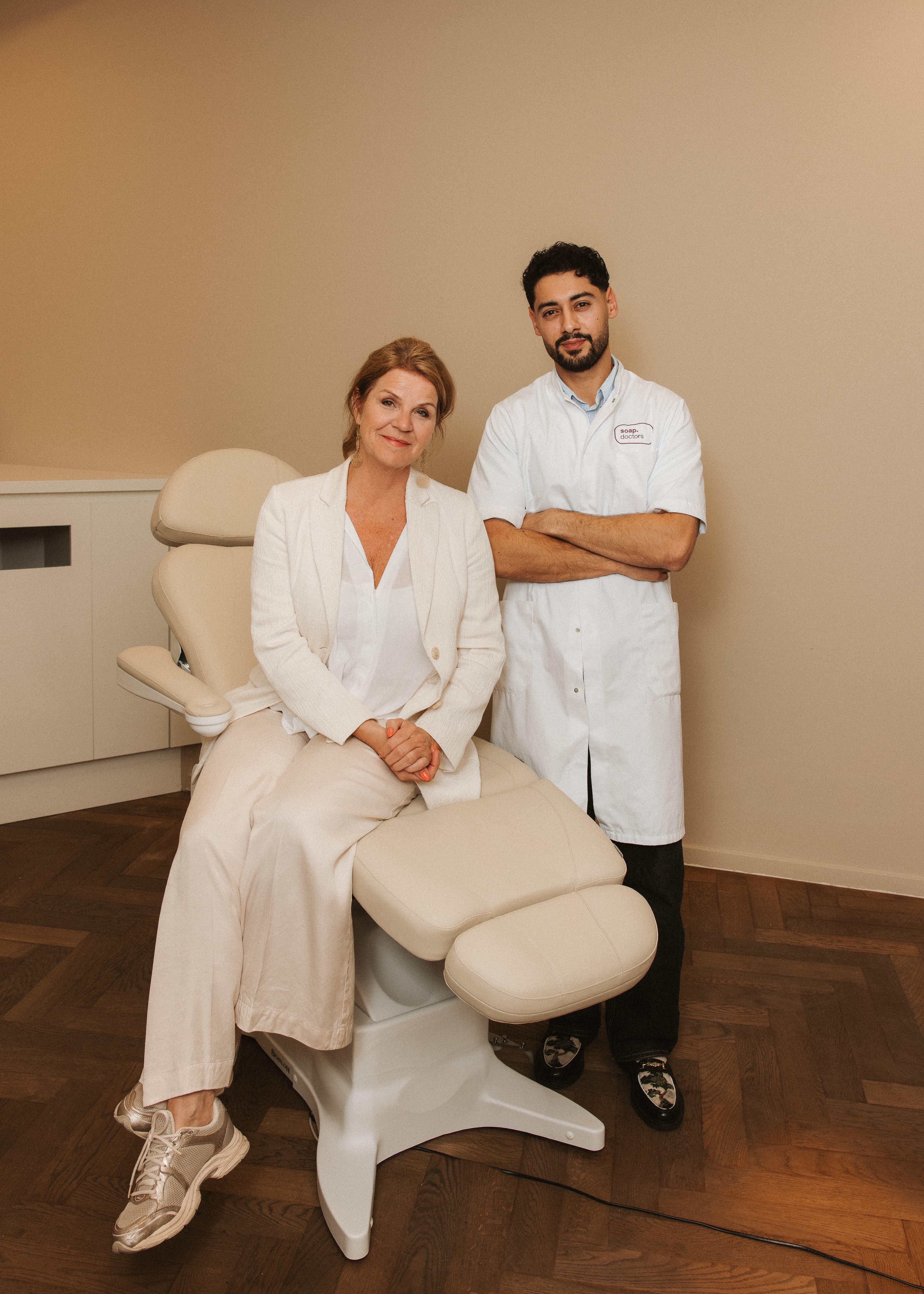 Een vrouw zit in een behandelstoel naast een man in witte jas met over elkaar geslagen armen in een behandelkamer
