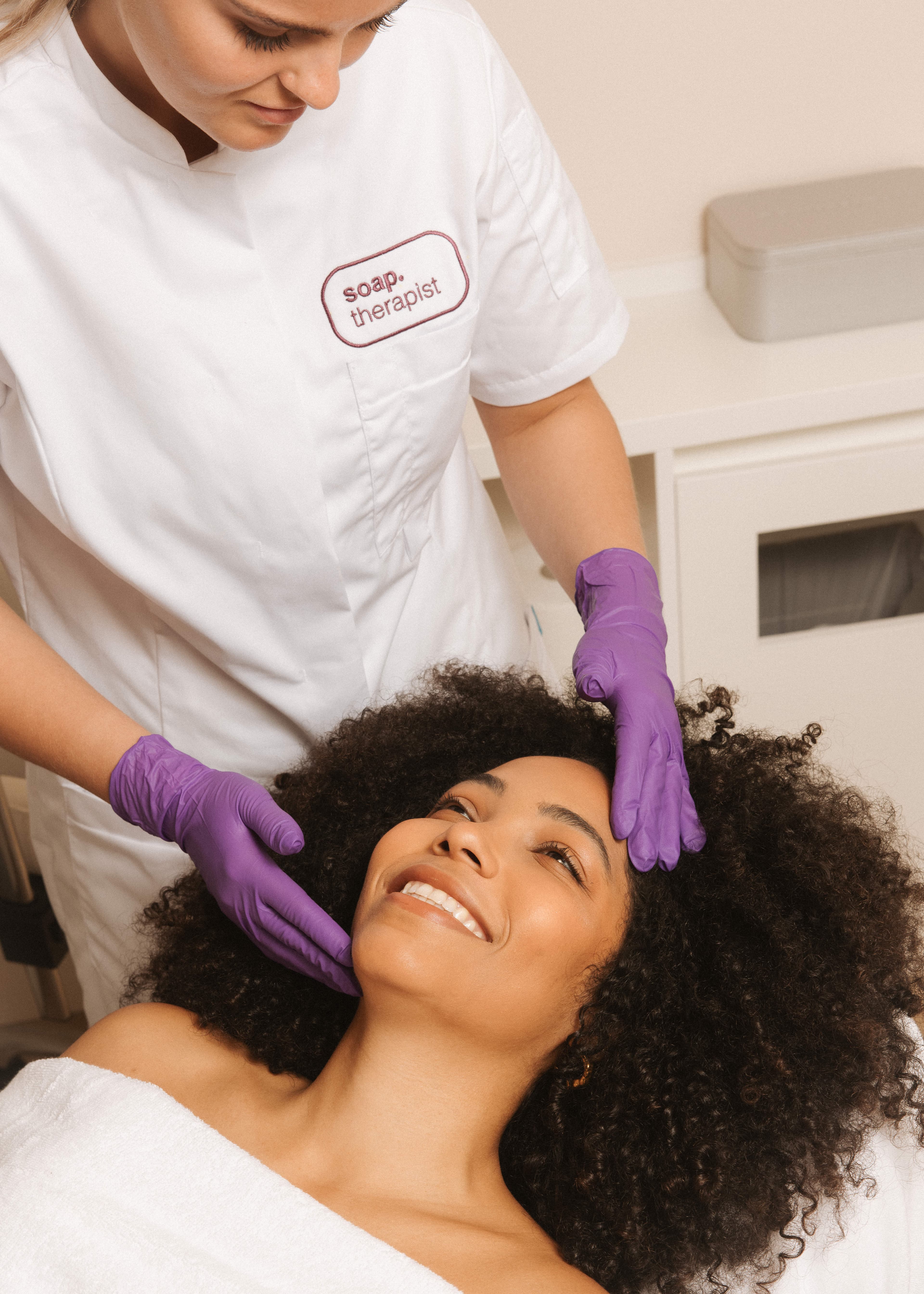 Therapist in a white coat and purple gloves gently massages a smiling client's face with curly hair, lying on a spa bed.