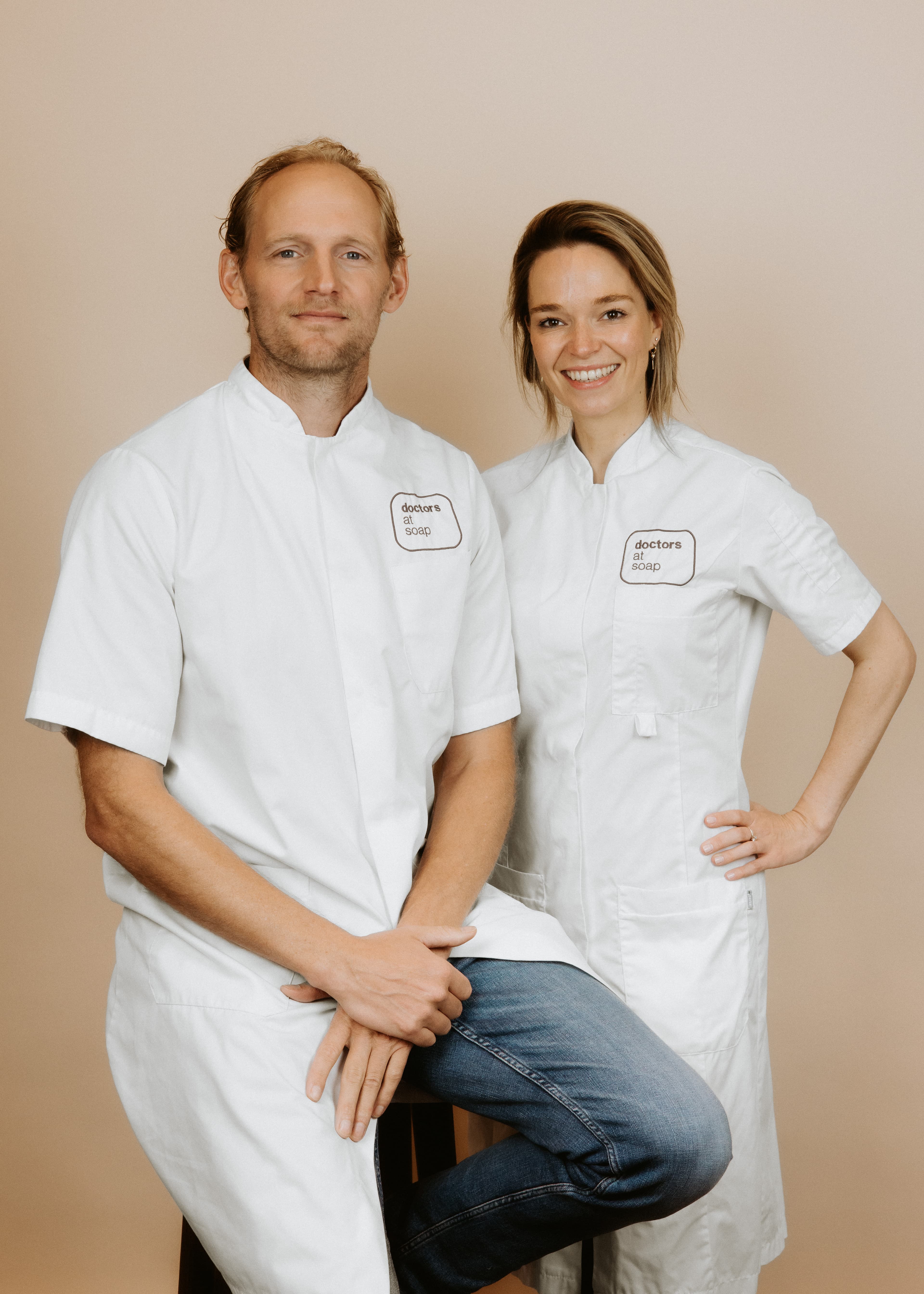 A man and woman in white uniforms, smiling and posing against a beige background. The man sits on a stool while the woman stands with one hand on her hip.