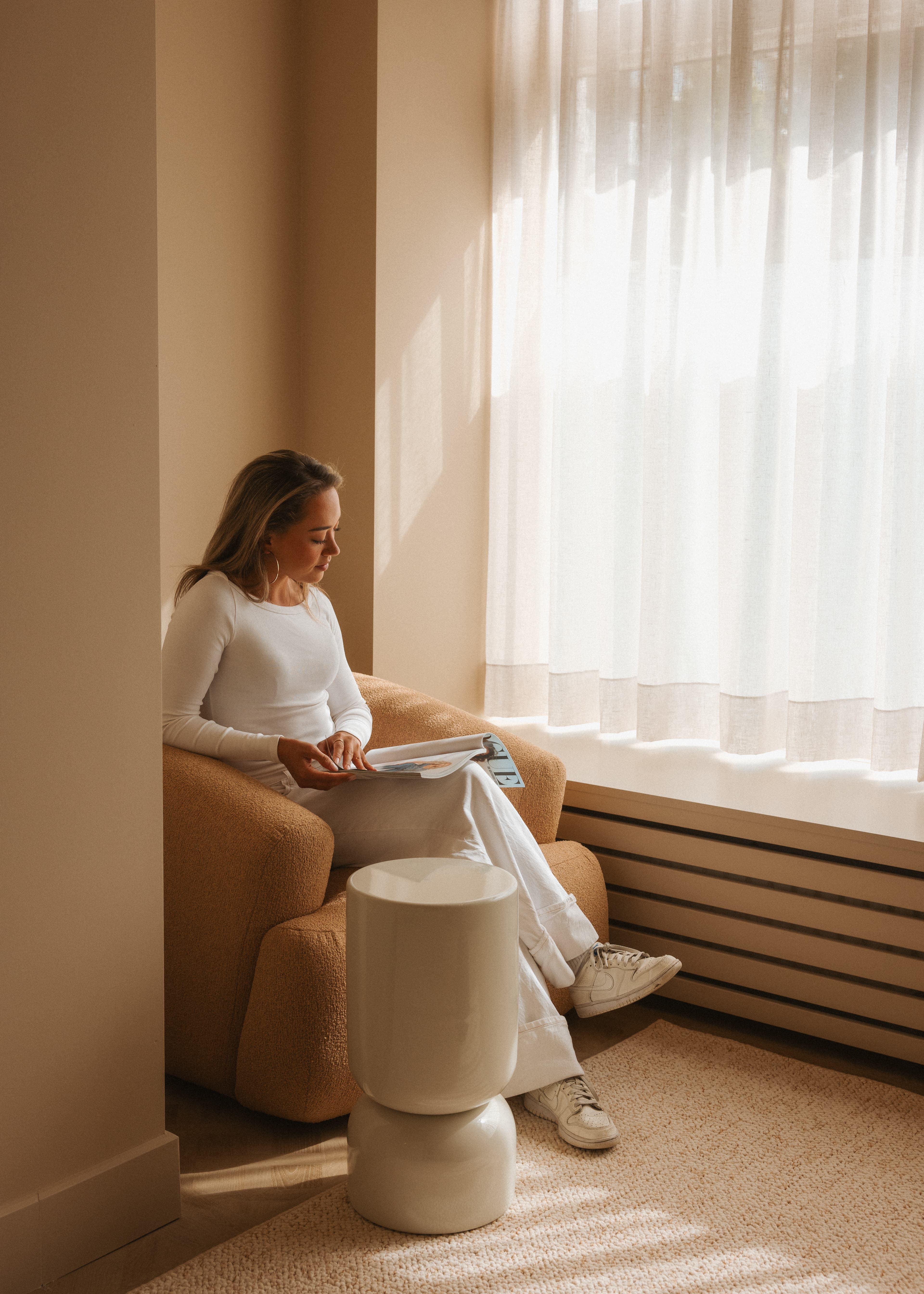 A woman in white clothes sits on a beige armchair by a sunlit window, reading a magazine. A small round table is in front of her.
