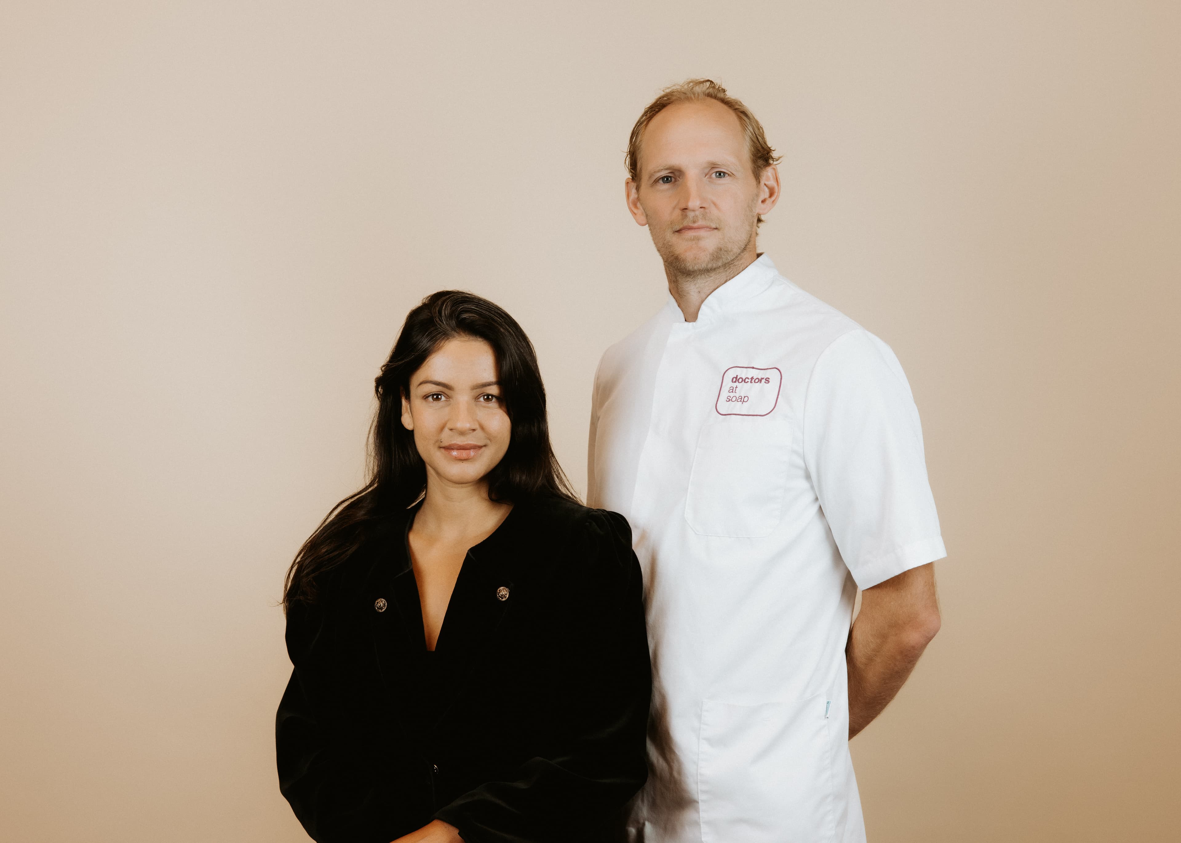 A woman in a black blouse and a man in a white chef's coat stand side by side against a plain background.