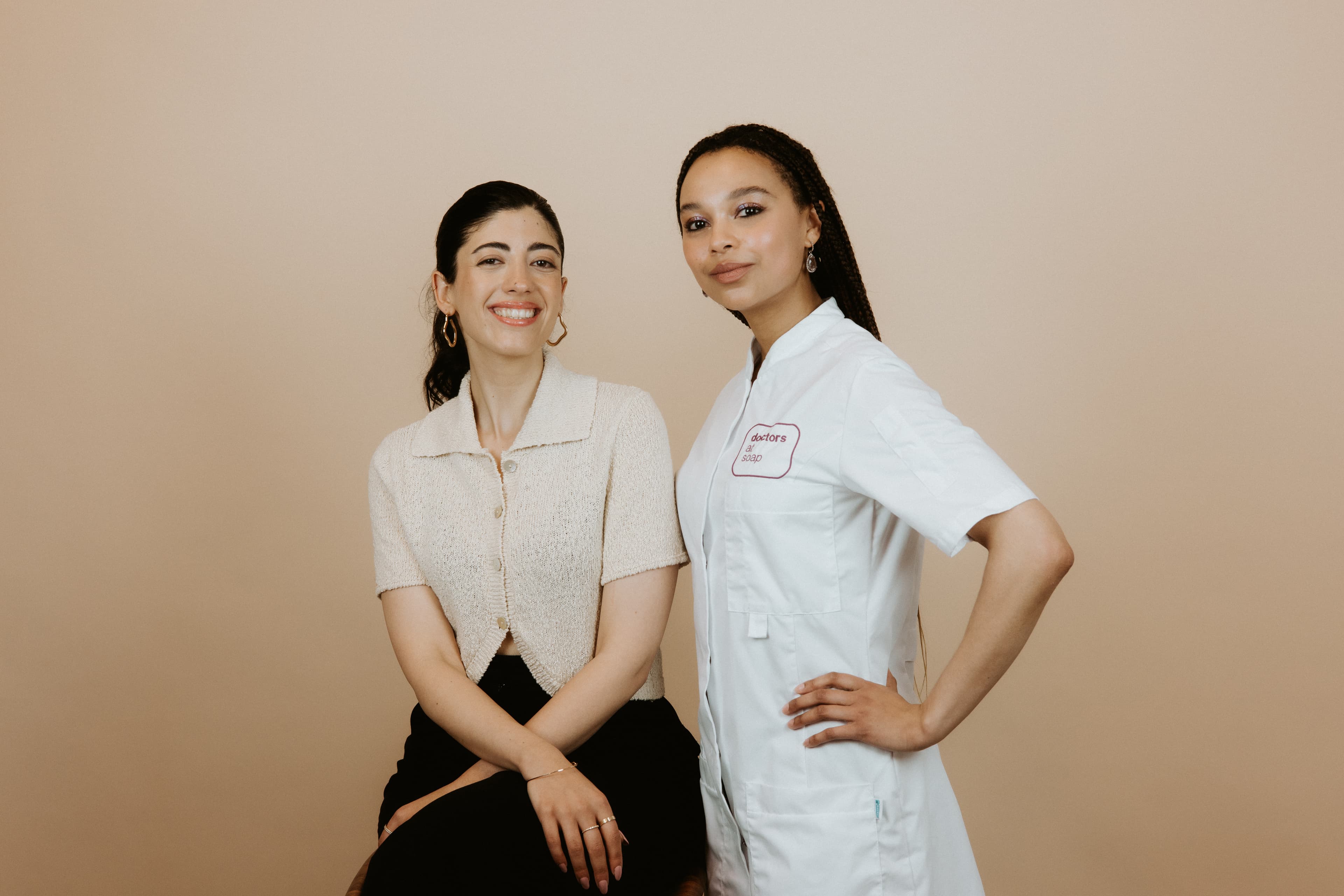 Two women pose together; one seated in a casual outfit, the other standing in a professional uniform against a beige background.