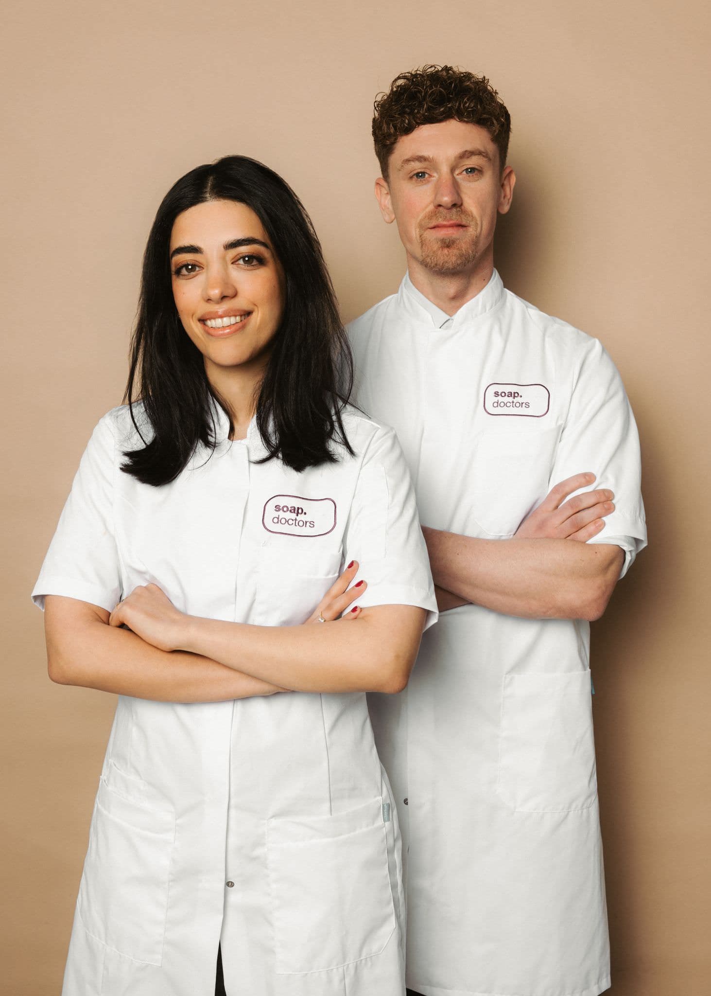 Two people in white lab coats labeled "Soap Doctors," standing confidently with arms crossed, smiling against a beige background.