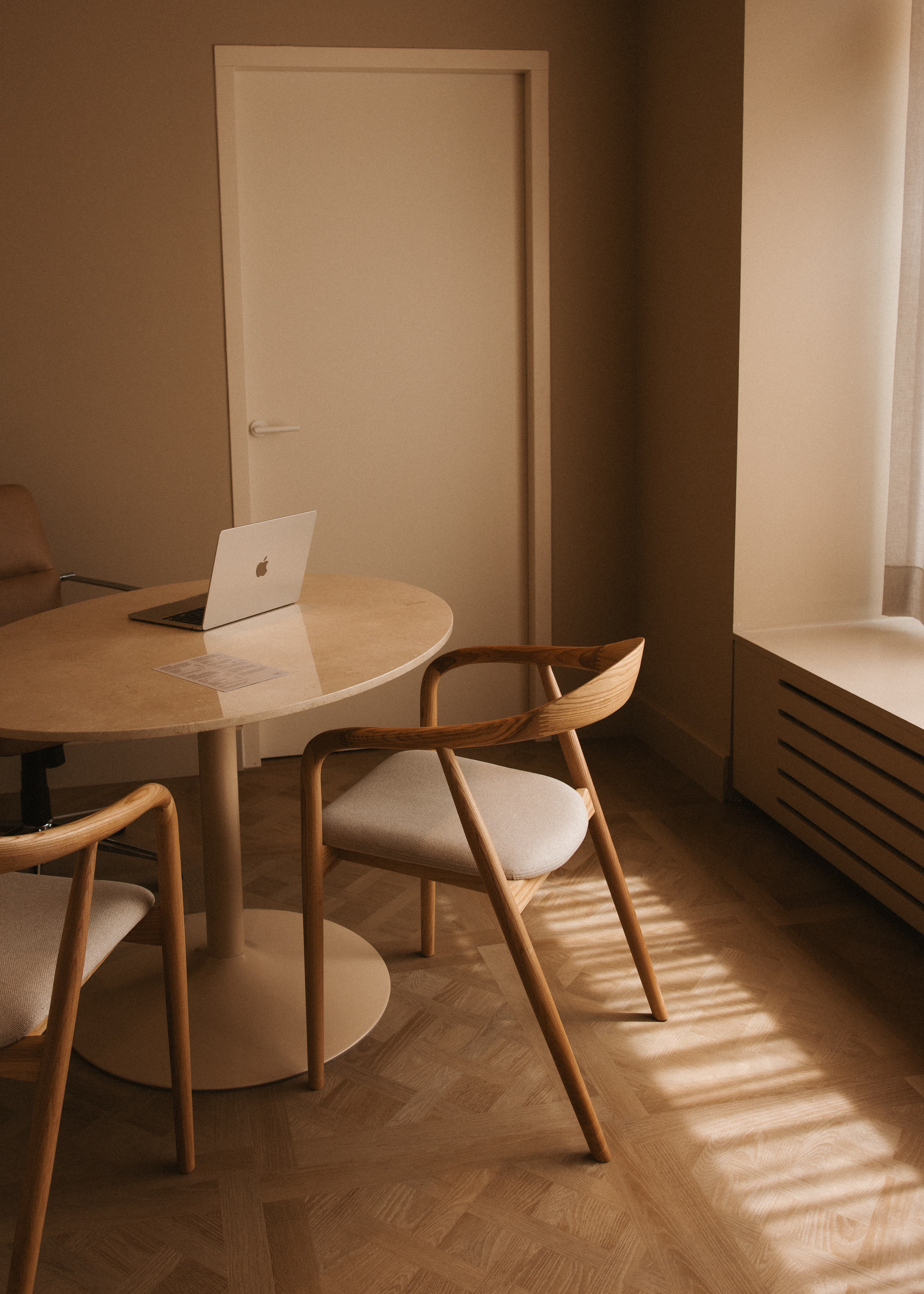 A minimalist office setting with a round wooden table, two chairs, a closed laptop, and warm sunlight streaming through a window.
