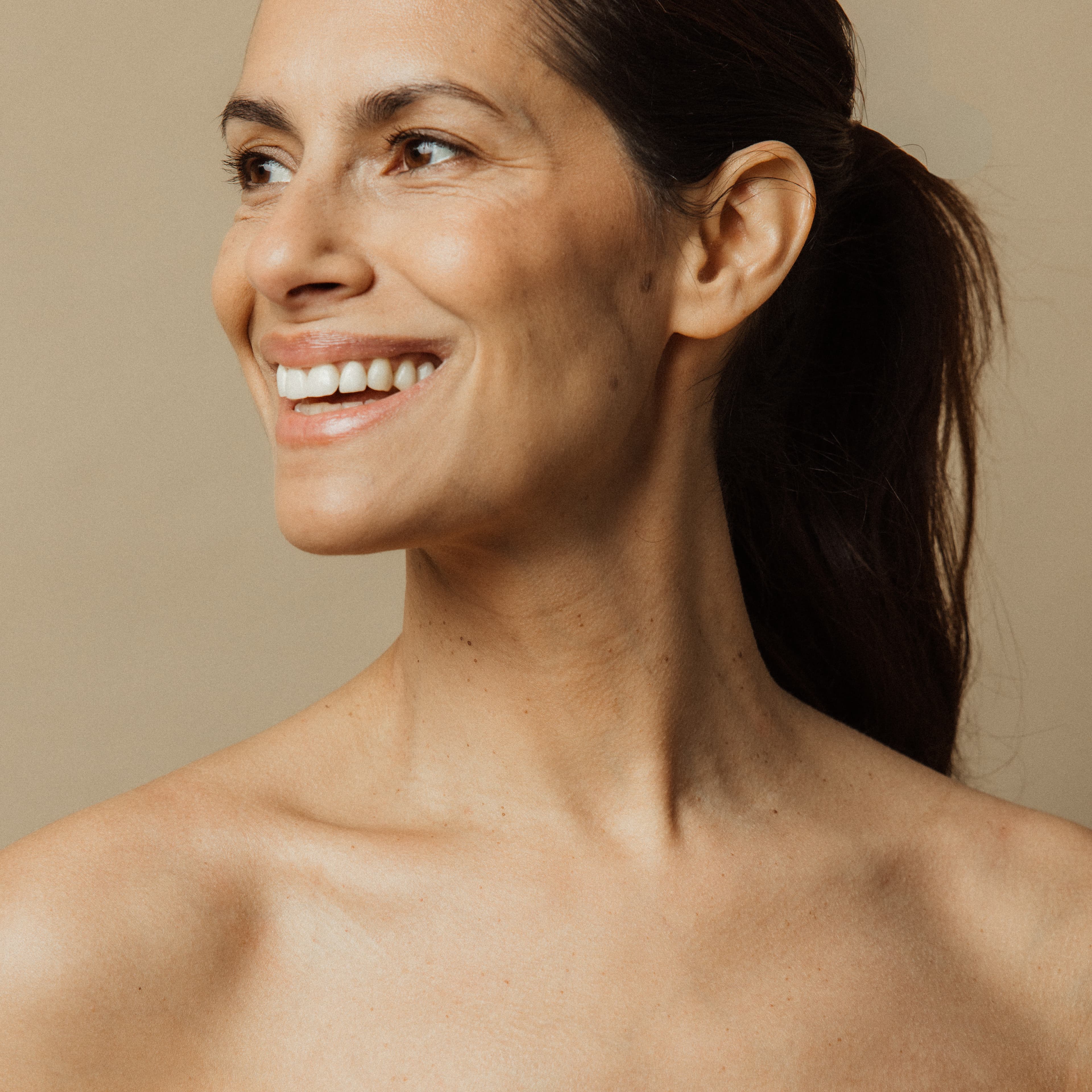 Smiling woman with dark hair in a ponytail, looking to the side against a neutral background.