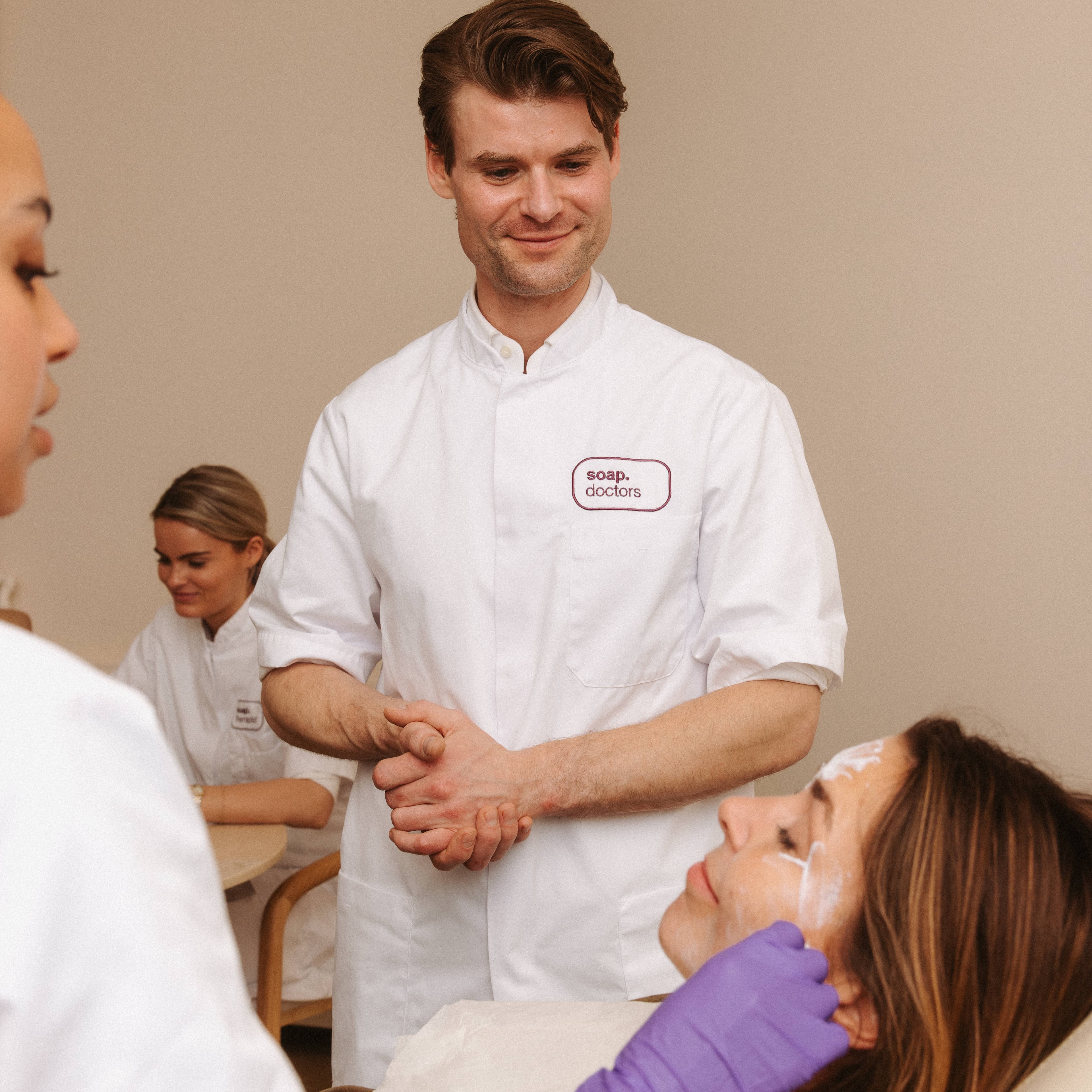 A man in a white coat observes a beauty treatment being applied to a woman's face by a gloved individual.