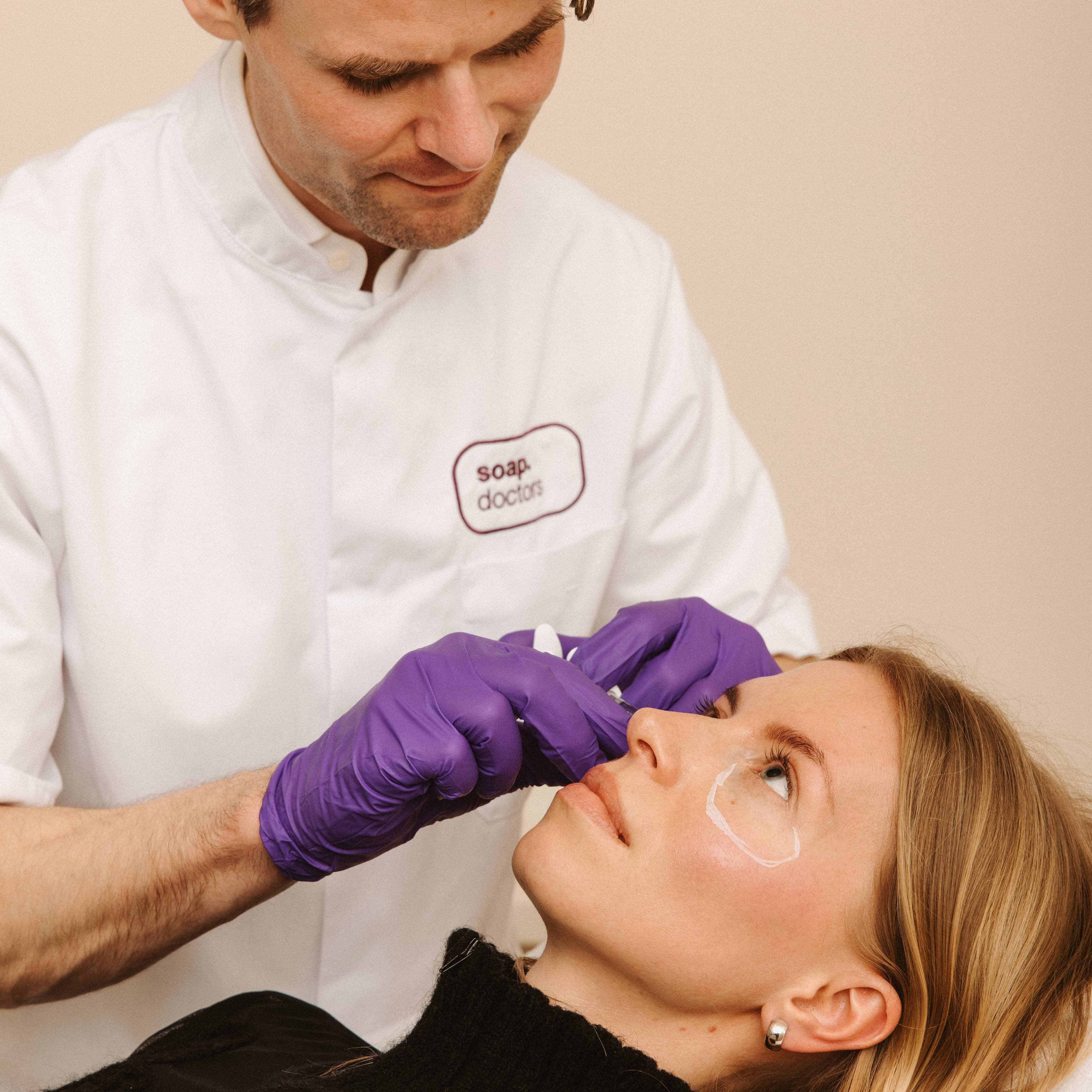 A professional in a white coat and purple gloves performs a cosmetic procedure on a woman lying down, with eye treatment patches applied.